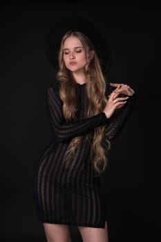 Portrait of shy young blonde looking down and crossing arms shyly at elbows. Model poses on black background. Blonde with long hair wears short black knitted see-through tight dress and felt hat