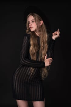 Portrait of shy young blonde looking away, shyly raising one arm bent at elbow, supporting it with other. Blonde with long hair wears short black tight knitted see-through dress, wide brimmed felt hat