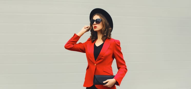 Stylish elegant woman posing in business suit, red blazer jacket, black round hat with handbag clutch