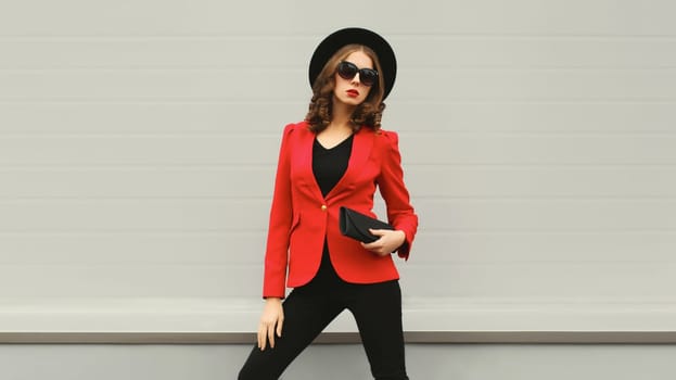 Stylish elegant woman posing in business suit, red blazer jacket, black round hat with handbag clutch