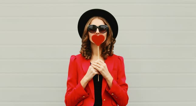 Beautiful stylish woman blowing kiss with sweet red heart shaped lollipop on stick in black round hat