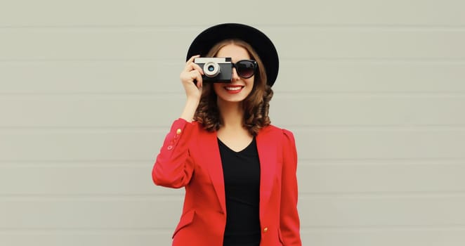 Beautiful stylish young woman photographer with film camera taking picture in black round hat, red blazer jacket