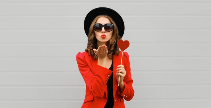 Beautiful stylish woman blowing kiss with sweet red heart shaped lollipop on stick in black round hat