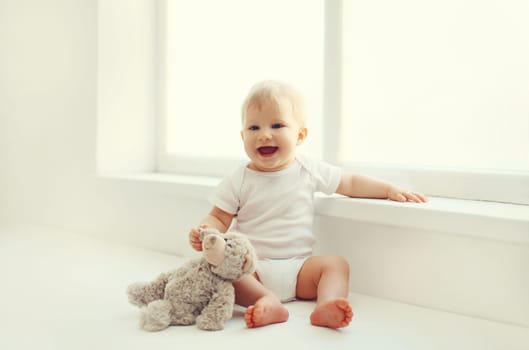 Happy cute little baby playing with teddy bear toy on the floor in white room at home