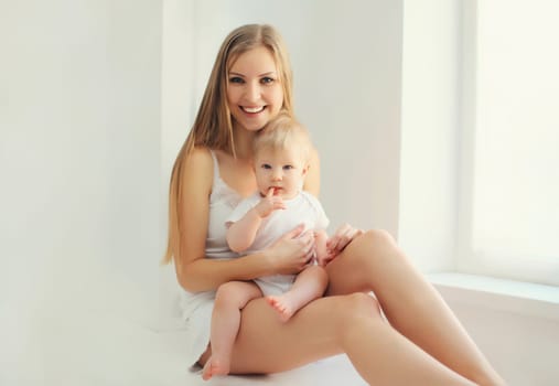 Happy smiling young mother playing with cute baby sitting together in white room at home