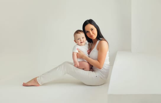 Happy young mother holding her cute baby sitting together in white room at home