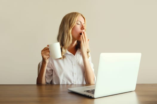 Tired overworked sleeping woman employee yawns, drinks coffee while working with laptop sitting at desk in office in morning