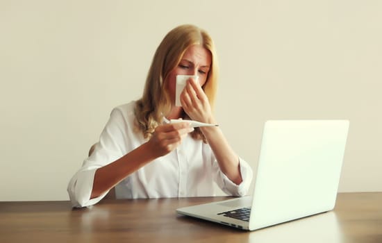Sick woman sneezing blow nose using tissue, measuring checking body temperature with thermometer working with laptop sitting at desk in office