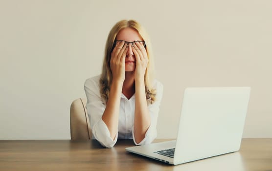 Tired overworked woman employee suffering from headache, rubbing her dry eyes, working with laptop sitting at desk in office