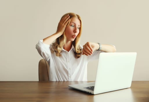 Shocked employee young woman working with laptop looks at watch, checks the time, fails to complete a project on deadline sitting at desk in office