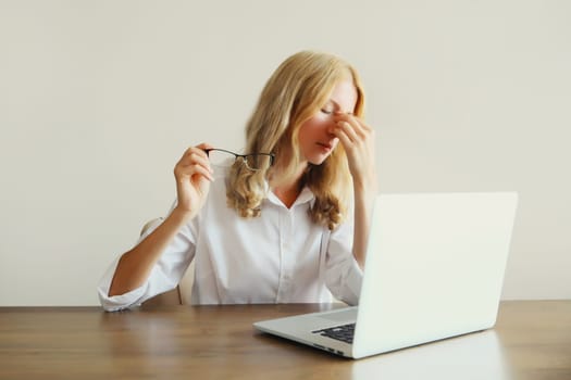 Tired overworked woman employee suffering from headache, rubbing her dry eyes, working with laptop sitting at desk in office