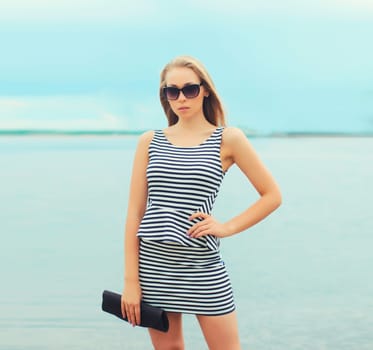 Portrait of beautiful blonde young woman posing in sunglasses, striped dress with handbag clutch on sea coast background