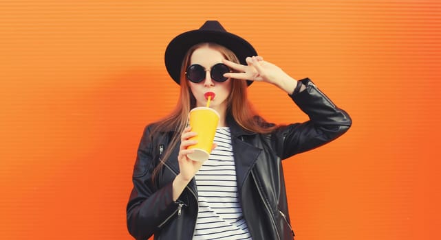 Portrait of stylish young woman drinking fresh juice posing in black rock style leather jacket, round hat on orange background