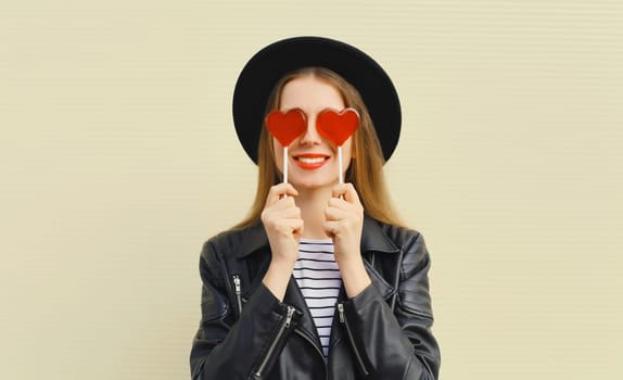 Portrait of happy cheerful young woman with sweet red heart shaped lollipop on stick in black round hat on studio background