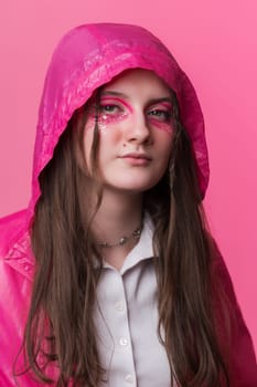 Portrait informal young adult woman with pink stage makeup with sequins and arrows painted on face, looking at camera. Female dressed in pink jacket with hooded over. Studio shot on colored background