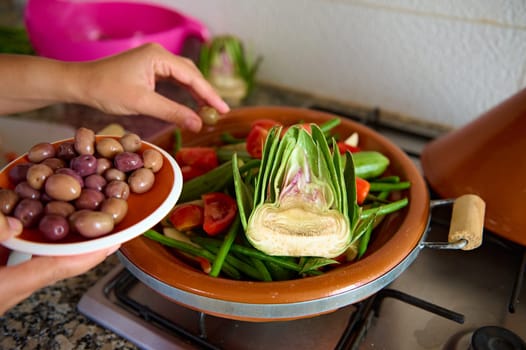Close-up housewife adding olives into a meal while cooking healthy vegetarian meal in tagine. Woman preparing and seasoning veggies at home