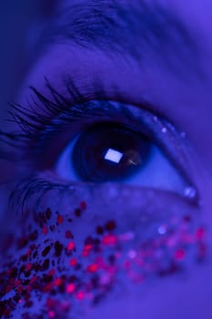 Extreme close-up view of female eye with shiny make-up with sparkles. Beauty night macro shot of female eye illuminated pink and blue color neon light. Selective focus. Part of photo series