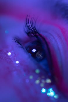 Extreme close-up view of female eye with shiny make-up with sparkles and arrows. Beauty night macro shot of female eye illuminated pink and blue color neon light. Selective focus, part of photo series