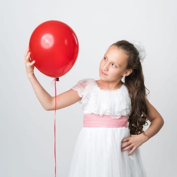 Portrait of girl in white dress holding one red balloon in hand and looking at balloon. Studio shot of Caucasian model 10 years old on white background. Part of photo series