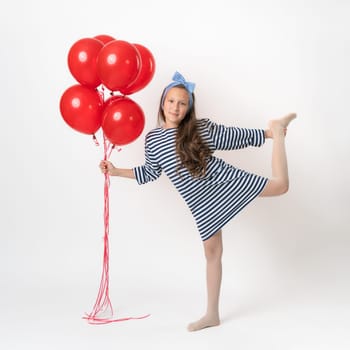 Active girl posing, holding bunch of red balloons in hand and looking at camera. Caucasian model 10 years old in white blue striped dress standing on one leg on white background. Part of photo series