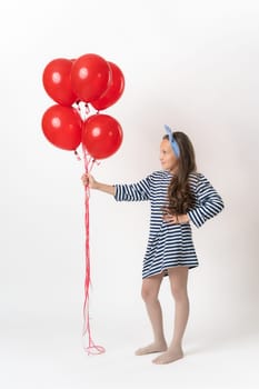 Girl in striped dress holding large bunch of red balloons on outstretched hand and looking at them. Young fashion model 10 years old standing full length on white background. Part of photo series