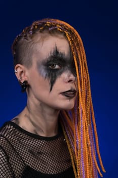 Dramatic portrait of young woman with creative horror black stage makeup painted on face and orange color without dangerous dreadlocks hairstyle. Studio shot on blue background. Part of photo series