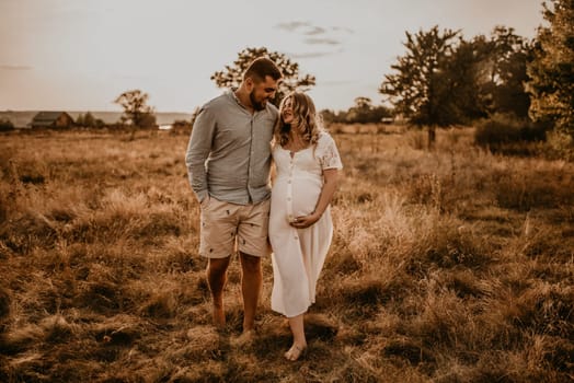 Happy family pregnant caucasian blonde woman with moles on face in white cotton dress walks with husband meadow summer. man in light natural clothes and shorts holds hand wife. trees in sunbeams