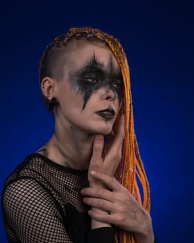 Cinematic portrait of beautiful young woman with orange color dreadlocks hairstyle and horror black stage make-up painted on face. Studio shot on blue background. Part of photo series