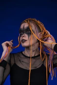 Cinematic portrait of young woman with horror black stage make-up painted on face and orange color without dangerous dreadlocks hairstyle. Studio shot on blue background. Part of photo series