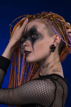 Beautiful adult female with orange color dreadlocks hairstyle and creative horror black stage makeup painted on face. Studio shot on blue background. Part of photo series