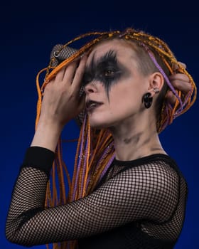 Dramatic portrait of adult female with horror black stage makeup painted on face and orange color without dangerous dreadlocks hairstyle. Studio shot on blue background. Part of photo series