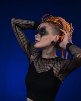 Female with orange color braids hairdo and horror black stage makeup painted on face. Studio shot on blue background. Part of photo series