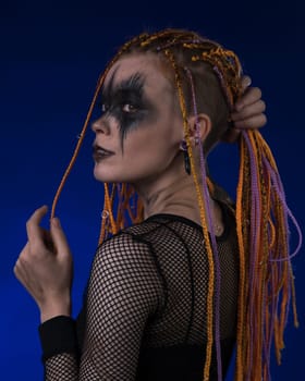 Dramatic portrait of young female with horror black stage makeup painted on face and orange color dreadlocks hairstyle. Studio shot on blue background. Part of photo series