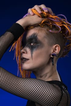 Headshot of young woman with horror black stage make up painted on face and orange color without dangerous dreadlocks hairstyle. Studio shot on blue background. Part of photo series