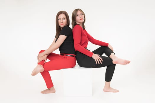 Woman mom and daughter sitting with their backs to each other on cube on white background. Both women crossed their legs and seriously looking over their shoulders at each other. Part of photo series