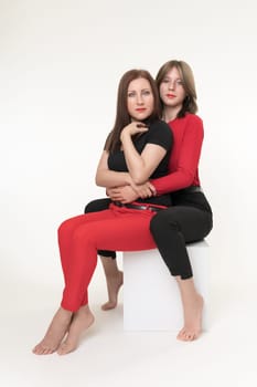Mother and daughter sitting on white cube box on white background. Daughter sits behind and hugging mom. Both females looking seriously at camera. Togetherness concept. Part of photo series