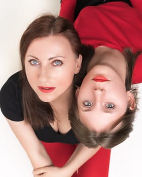 High angle shot of mother and teenager daughter sitting with their backs to each other, put their heads on each other's shoulders. Both females looking up at camera. Studio shot, part of photo series