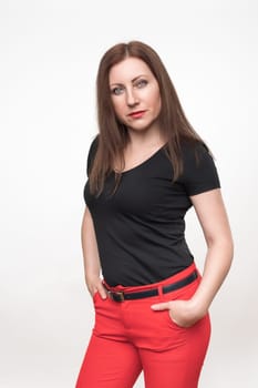 Portrait of Caucasian ethnicity woman forty years old on white background. Adult woman dressed in black t-shirt and red pants holds hands in pockets and looking intently at camera.