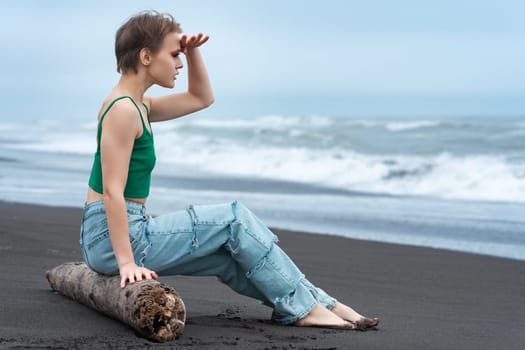 Hipster woman sitting on log on black sandy beach of Pacific Ocean, raised her hand to forehead and looking at view at breaking sea waves. Blonde woman with short hair wearing green top and blue jeans