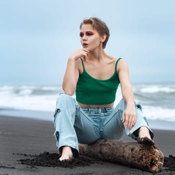Blonde woman with short hair and bright makeup is sitting on log on beach, looking away with hand resting on chin. Hipster woman is enjoying serene and peaceful scene on sandy beach of Pacific Ocean