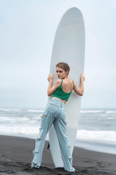 Rear view of woman surfer standing on sandy beach, holding surfboard vertically and looking over shoulder at camera. Fashion model wearing casual clothing, posing on background of breaking ocean waves