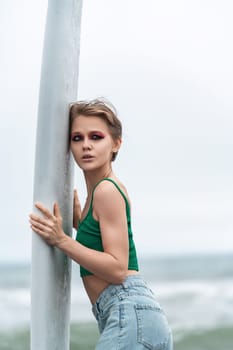 Portrait of sensual woman holding surfboard with both hands, presses against it and looking away. Caucasian fashion model with bright make-up and short hair wearing casual clothing - top, blue jeans