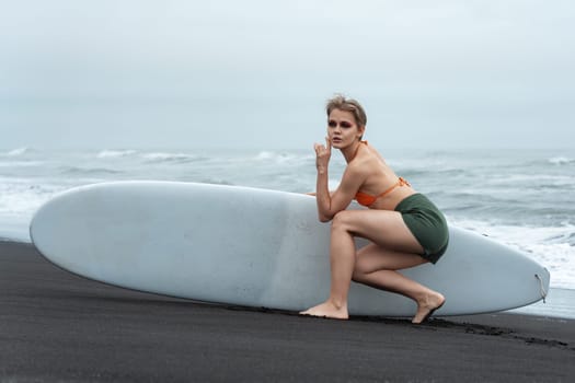 Female sports fashion model holds surfboard and sits in front of it, looking thoughtfully into distance against background of ocean waves. Beautiful woman is wearing bikini top and shorts