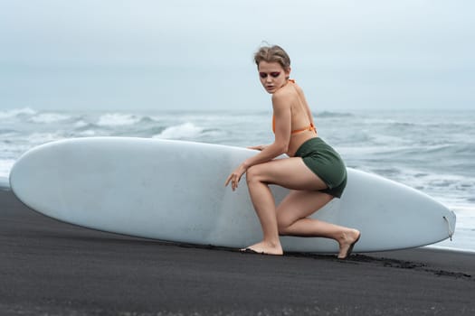 Female sports fashion model holding surfboard and sits in front of it, thoughtfully looking down against backdrop of sea waves. Beautiful woman surfer is wearing bikini top and shorts