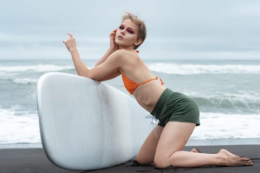 Female surfer exudes confidence as she gracefully kneels on black sand beach, holding surfboard. Alluring pose, complemented by bikini top and shorts, showcases athletic prowess and self-assurance.