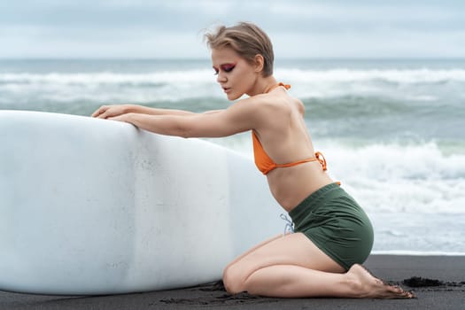 Female surfer, with her eyes closed, is kneeling and holding her surfboard during summer vacation. Sportswoman is wearing bikini top and shorts, and appears to exude sensuality