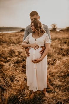 husband hugs pregnant wife keep his hands on big round tummy. Happy family resting in nature hugs kisses in summer at sunset. future mother Caucasian woman in white cotton dress father in shorts