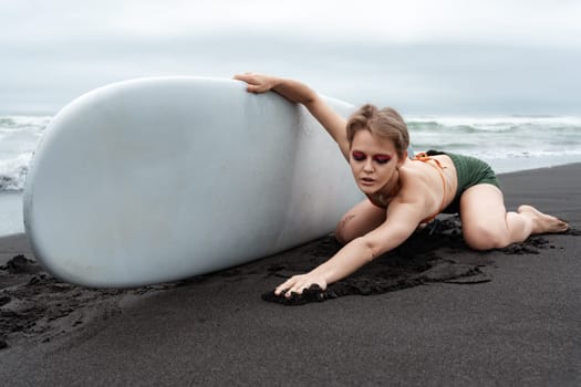 Female surfer looks stunning as she kneels on black sand beach, stretching forward with her back arched while holding surfboard, looking down. Fashion model is rocking stylish bikini top and shorts