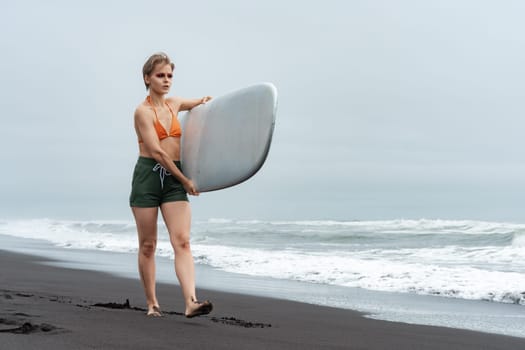 Sportswoman surfer in bikini top, shorts and barefoot walking on black sand beach and carrying white surfboard against background of sea waves during summer vacation. Concepts of sports activities