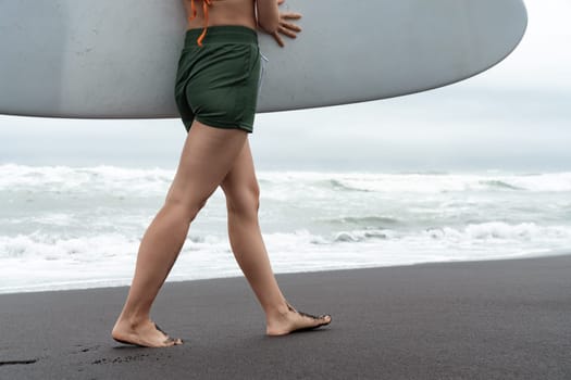 Surfer woman carrying surfboard walking along beach with sea waves in background. Side view of sensuality female legs and buttocks is an athletic figure. Concept of extreme sports